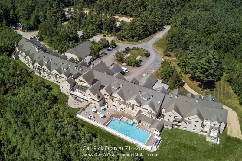 Aerial view of a large, multi-story hotel surrounded by dense forest. The building has a grey roof, numerous windows, and an outdoor pool area with blue water and poolside furniture. Vehicles are parked in the front area, and winding roads provide access.