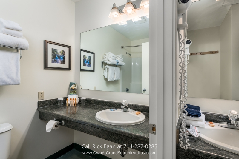 A hotel bathroom with a large mirror above a dark marble countertop with a sink. White towels are folded and stacked on towel racks. Toiletries are arranged near the sink. Wall art featuring a nature scene and an overhead light fixture are visible.