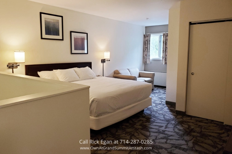 A hotel room with a queen-sized bed adorned with white linens, flanked by wall-mounted lamps, and two framed pictures above the headboard. There's a beige couch near a curtained window, and a closed sliding door on the right. The room features patterned carpet flooring.