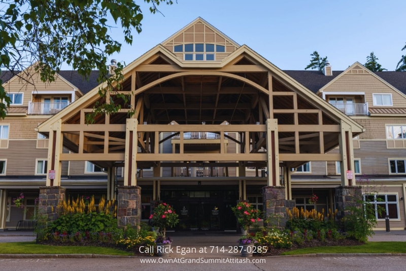 A large lodge-style building with beige wooden siding and stone accents, featuring a prominent A-frame entrance. Mature trees and a bright blue sky frame the background. The entrance is flanked by colorful flowers and decorative potted plants.