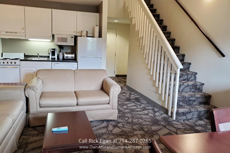 A cozy living area with a beige couch, a wooden coffee table, and a staircase leading upstairs. The kitchenette features white cabinets, a fridge, a microwave, and a stove. A TV remote is on the coffee table. Walls and flooring are light-colored with a patterned carpet.