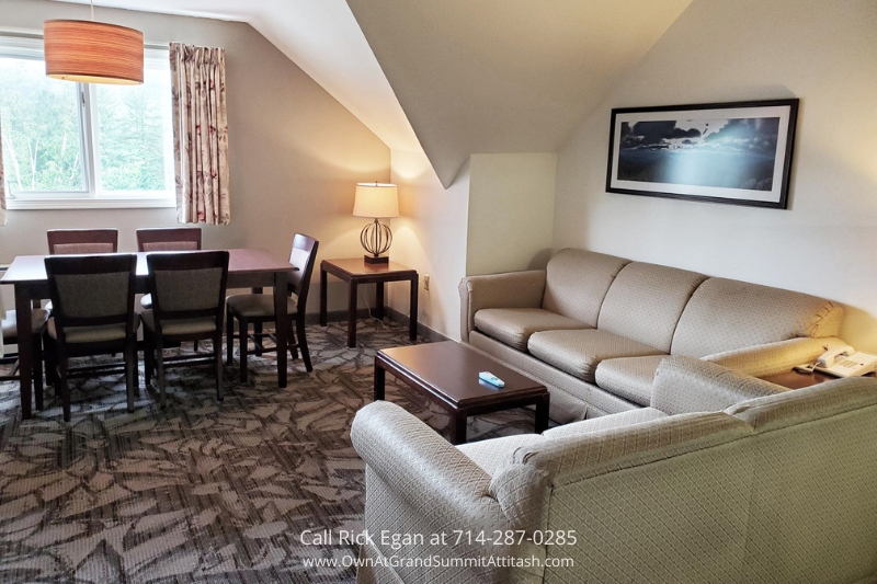 A cozy living area with a beige sectional sofa, a wooden coffee table, and a floor lamp. Adjacent is a dining area with a rectangular table and six chairs. A large window with patterned curtains lets in natural light, and a framed photograph hangs on the wall.