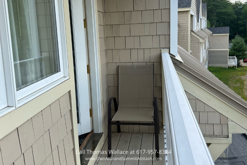 A small balcony with beige siding features a single patio chair. The door to the balcony is open, providing a view of adjacent townhouses and greenery in the background. The balcony has a white railing that partially extends out of the frame.