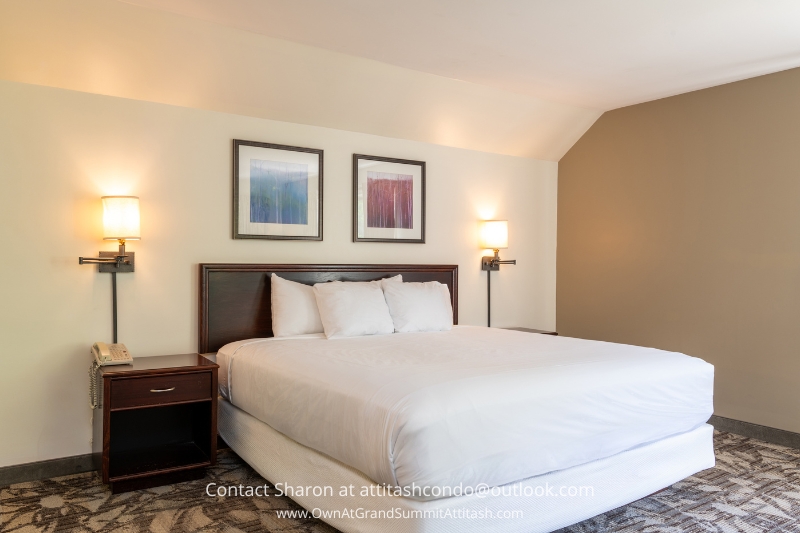 A neatly made bed with white linens sits between two wall-mounted lamps in a hotel room. A side table with a phone and a drawer is on the left side of the bed. Two framed abstract paintings hang above the headboard. The carpet has a geometric pattern.