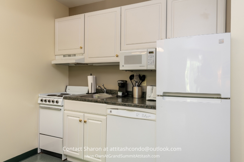 A small kitchen with white cabinets, a white refrigerator, an oven, and a microwave. The countertop holds a knife block, cooking utensils in a holder, a dish rack, and a coffee maker. The kitchen has a minimalist design with beige walls.