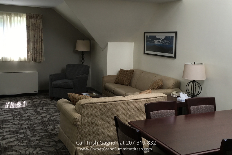 Open-concept living room in a unit at the Grand Summit Hotel, featuring a comfortable beige sofa, armchair, and coffee table. The room has soft lighting from floor lamps, a large window with curtains, and a stylish patterned carpet, making it a perfect space for relaxation.