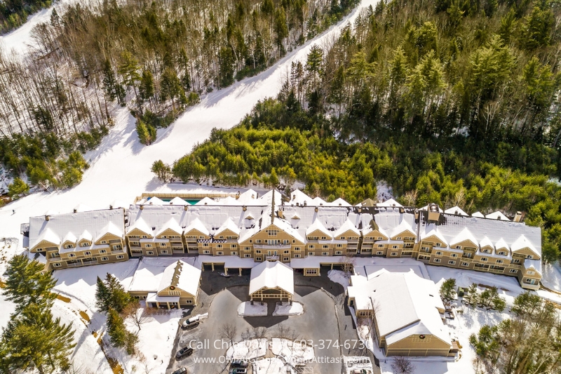 A stunning winter aerial view of the Grand Summit Hotel, showcasing its snow-covered roof, surrounded by dense evergreen forests and adjacent ski trails in Bartlett, NH.