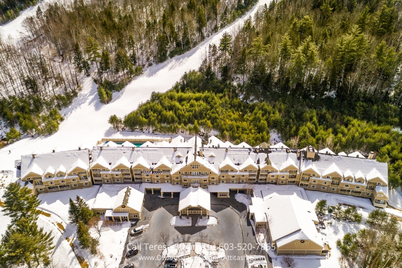 A picturesque aerial view of the Grand Summit Hotel covered in snow, surrounded by evergreen forests and nearby ski trails, capturing the charm of winter resort living in Bartlett, NH.