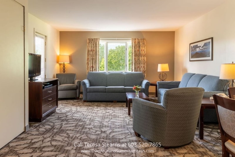 Bright and spacious living room in a unit at the Grand Summit Hotel, featuring comfortable gray sofas, a coffee table, a large window with a green outdoor view, and warm ambient lighting.