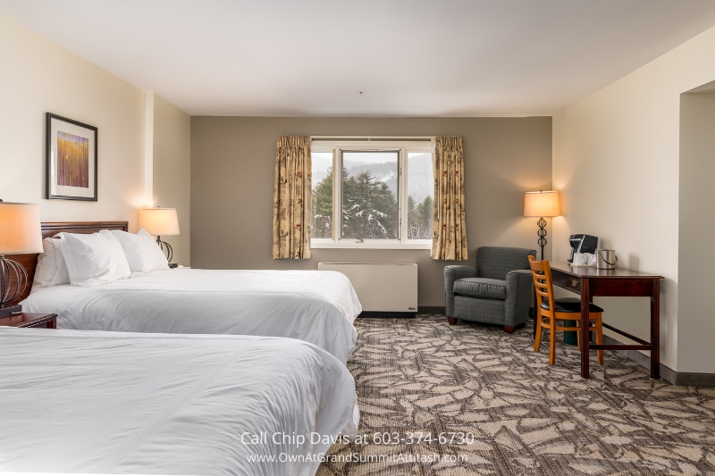 A serene bedroom at the Grand Summit Hotel, featuring two plush queen beds, a cozy armchair, a work desk, and a large window overlooking the picturesque scenery of Bartlett, NH.